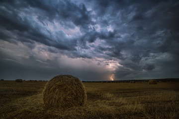 Rags of hay on the field and lightning. A dark night photo, a stormy sky and sparkling lightning on the field. Farm landscape.
