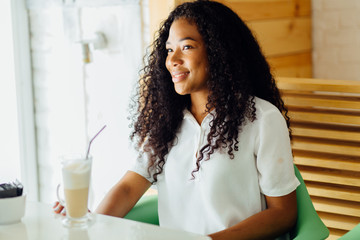 Beautiful young african woman sitting in a cafe, drinking coffee latte and and smiling at camera.