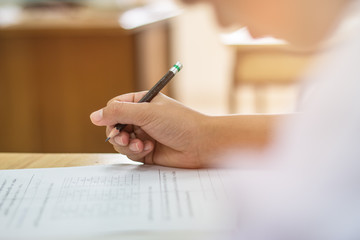Blurred of Asian boy students hand holding pencil writing fill in Exams paper sheet or test papers on wood desk table with student uniform in exam class room, education concept
