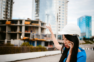 Asian woman in a blue dress and white protective helmet. engineer supervises inspection and control of works at construction site. Business telephone conversations. Communication via smartphone