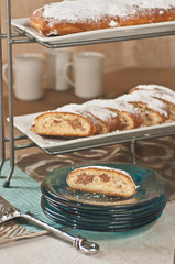 Slice of almond fig coffee cake on green, clear, round, stack of dishes, power sugar, artisan serving wedge and a metal stand, rectangular, white plates of sliced coffee cake 