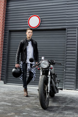 Beautiful young rider guy in black biker jacket and boots go to his classic style cafe racer motorcycle industrial gates as background. Bike custom made in vintage garage. Brutal fun urban lifestyle.