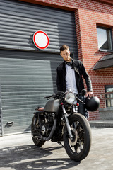 Handsome young rider man in black biker jacket and jeans go to his classic style cafe racer motorcycle industrial gates as background. Bike custom made in vintage garage. Brutal fun urban lifestyle.