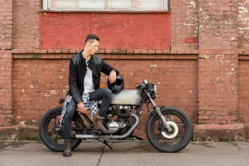 Fototapeta na wymiar Handsome rider biker guy in black leather jacket, boots and style jeans sit on classic style cafe racer motorcycle. Bike custom made in vintage garage. Brutal fun urban lifestyle. Outdoor portrait.
