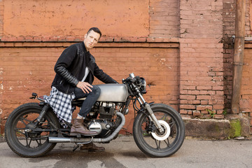 Plakat Handsome rider biker guy in leather jacket sit on classic style cafe racer motorcycle. Side view. Horizontal photo. Bike custom made in vintage garage. Brutal fun urban lifestyle. Outdoor portrait.