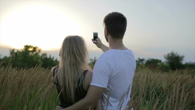 A loving couple, Guy with a beautiful young woman do selfie on the field at the sunset or sunrise