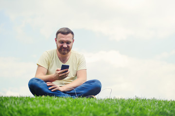 Naklejka na ściany i meble Nature and technology. A man with a beard lies on a green grass with a smartphone in his hands is charging power bank.
