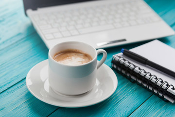 Coffee cup, notebook and laptop on the wooden turquoise background.  Selective focus on coffee cup