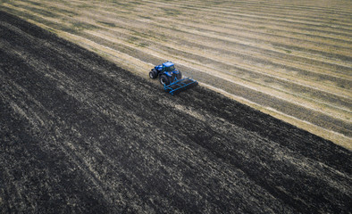 The white tractor plows the field against the backdrop of the black earth, and behind it birds fly and collect food. Aeril view. Agricultural machinery works in the field of spring planting. Plowing