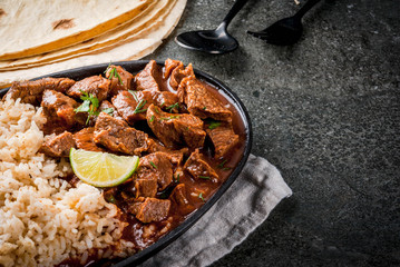 Mexican and American traditional food. Stew beef with tomatoes, spices, pepper - Chile Colorado. With boiled rice. lime, tortillas.  Copy space. On black stone table