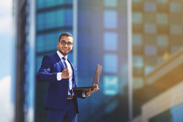 Competent business expert. Confident young handsome man in suit holding laptop and smiling while standing against office building