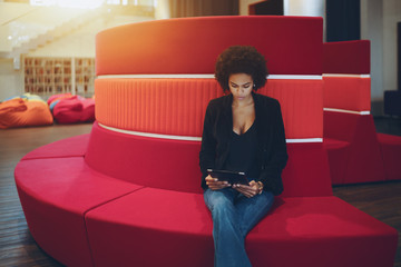 Young serious afro american girl with curly hair is sitting on red curved sofa in chillout office...
