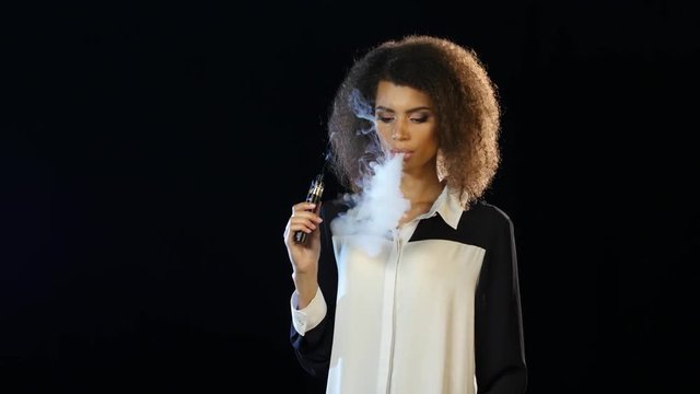 Girl of african american appearance smokes an e-cigarette. Black background