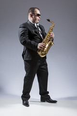 Fototapeta premium Music Ideas and Concepts. Full Length Portrait of Caucasian Saxophone Player in Sunglasses Playing the Instrument in Studio Environment.