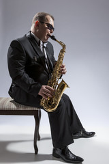 Fototapeta na wymiar Music Concepts. Portrait of Mature Relaxed and Thoughful Caucasian Saxophone Player in Sunglasses Playing the Saxophone While Sitting on Chair in Studio Environment.