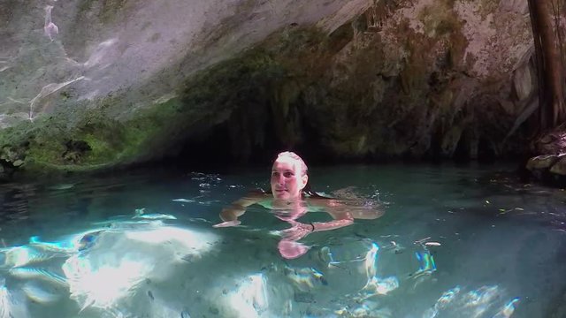 CLOSE UP, SLOW MOTION: Beautiful young woman swimming in fresh river water inside the famous Grand Cenote cave illuminated with summer sun. Tourist girl bathing and exploring underground karst cavern