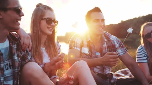 Young people having the picnic by the riverside, laughing happily, eating fried marshmallows and drinking beer. Having a little party. Friendship goals, summertime evenings. Positive atmosphere.