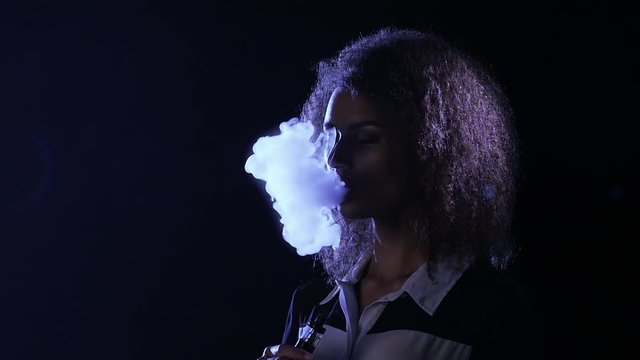 African american girl smokes an vaping in an empty room. Black background. Slow motion