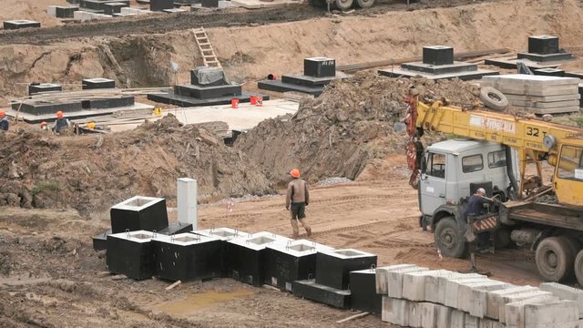Builder dressed in orange helmet walks next to foundation blocks. Mobile construction crane on wheels