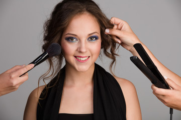 Many hands help young smiling woman with evening make-up on gray background in studio in studio