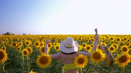 Happy woman in sunflower field playing on ukulele and singing. Summer girl in cowboy straw hat. Caucasian young lady jumping and have fun with raised hands. Slow motion. Little guitar. music concept.