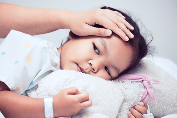 Fototapeta na wymiar Sick asian child girl is lying in the bed and mother hand touch her forehead with love in the hospital