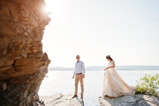 Fabulous wedding couple standing on the cliff next to a huge rock with a stunning view of lake in the background.