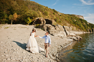 Amazing young couple holding hands on the lake shore on their sunny wedding day.