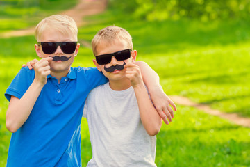 Two blond boys with fake mustaches on their faces and in sunglasses outdoors