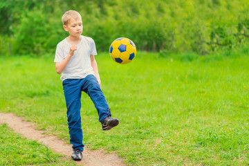 Blond boy in a white T-shirt walking along the path and kicking a yellow soccer ball. Toned