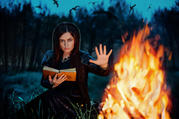 A young witch reading conjure from book at the fire at night in the forest. Priestess of fire....