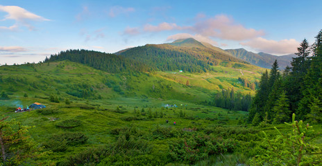 Tents and people in green lush valley against mountain tops covered with several clouds and forest, spruce trees. Warm summer evening. Panorama of Marmarosh, Carpathian mountains, Ukraine