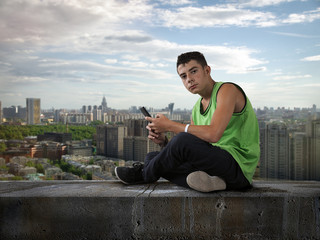Fototapeta na wymiar Young man with mobile phone on the roof. Teenager sits on top. Panorama of the city