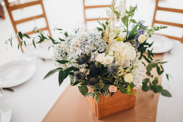 In the wedding banquet area there are wooden tables with tablecloths, on tables there are compositions of flowers and greens, candles, cutlery, on plates lie napkins with name cards