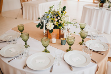 In the wedding banquet area there are wooden tables with tablecloths, on tables there are compositions of flowers and greens, candles, cutlery, on plates lie napkins with name cards