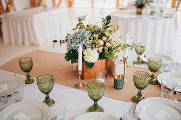 In the wedding banquet area there are wooden tables with tablecloths, on tables there are compositions of flowers and greens, candles, cutlery, on plates lie napkins with name cards