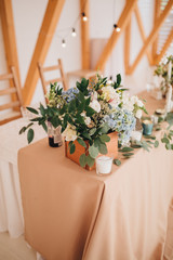 Fototapeta na wymiar In the wedding banquet area there are wooden tables with tablecloths, on tables there are compositions of flowers and greens, candles, cutlery, on plates lie napkins with name cards