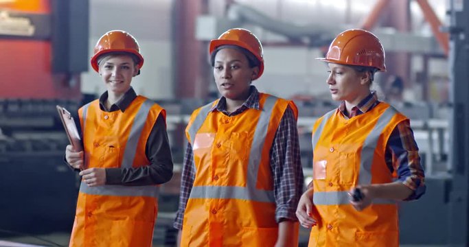 Slow motion shot of African female engineer walking with two colleagues, talking with them and showing something in factory building