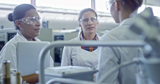Slowmo of black female engineer in lab coat holding laptop, smiling and talking with colleagues in industrial factory