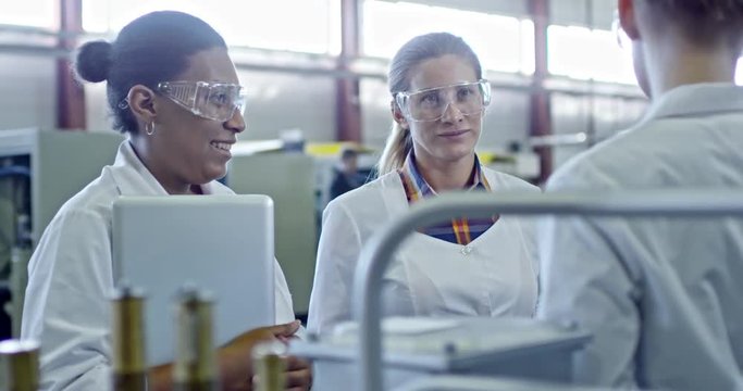 Slowmo of black female engineer in lab coat and protective eyeglasses standing with laptop and chatting with colleagues