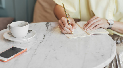 Hands of cropped unrecognisable woman writing in her notebook on coffee table.