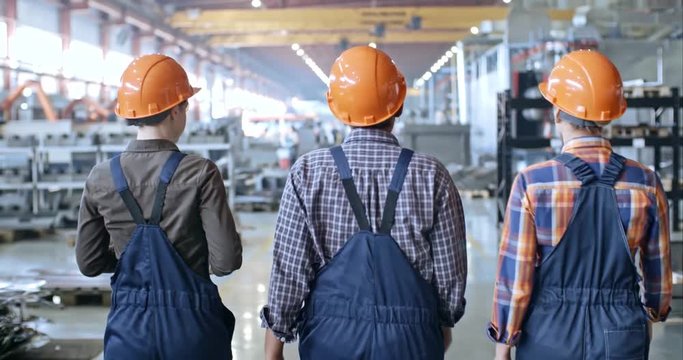 Rear view of three female workers in hardhats walking though factory and discussing something