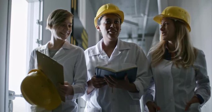 Three female engineers in hardhats and lab coats walking though corridor towards the camera; colleagues laughing while listening to African woman reading something in organizer