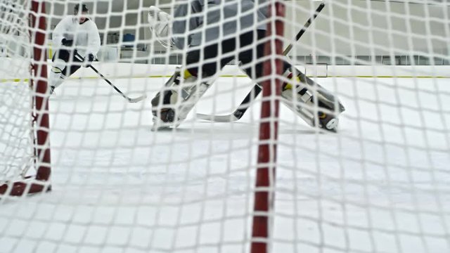 Slow motion shot behind net of hockey goalie tracking puck and guarding net, then performing butterfly technique and missing goal shot by forward in white uniform
