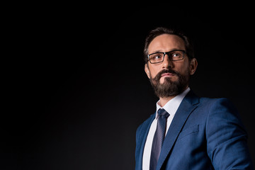 portrait of confident bearded mature businessman in eyeglasses looking away isolated on black