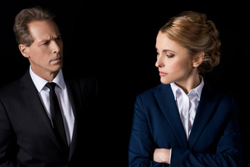 serious middle aged businesswoman standing with crossed arms and looking at colleague isolated on black