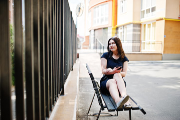 Brunette girl at black dress, sunglasses sitting on bench, listening music from headphones phone, and posing at street of city.