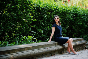 Brunette business girl at black dress on sunglasses sitting at border against bushes, posing at street of city.