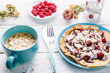 Homemade pancakes or Russian pancakes with chocolate sauce, whipped cream and raspberries on a plate on a white wooden background.