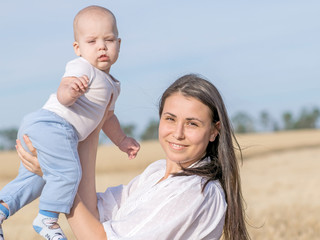 Young happy mother playing with her adorable newborn son in wheat field at summer day